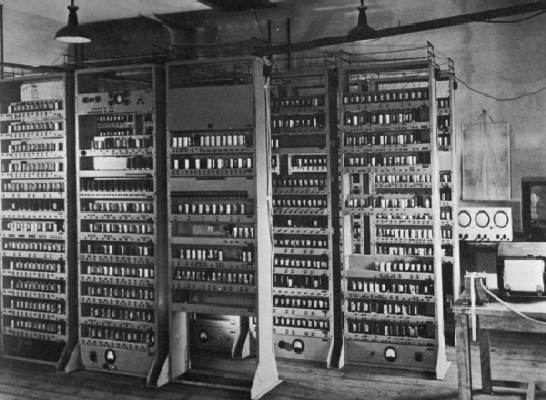 "A black and white photograph shows multiple tall vertical racks of the EDSAC computer system densely populated with vacuum tubes and electronic components. The image captures the large-scale physical architecture of early stored-program computers within a laboratory setting."